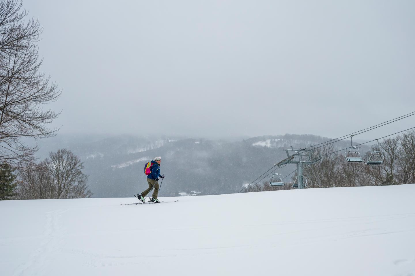 Skier on snowy slope under gray sky, with distant mountains and lift.