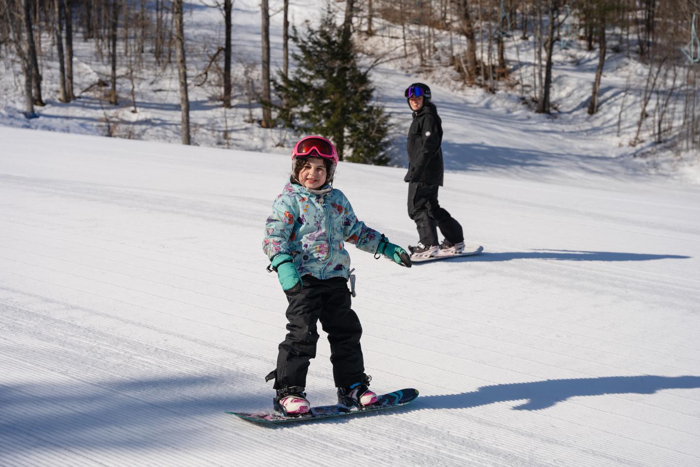 Child snowboards on a snowy slope, adult in background.