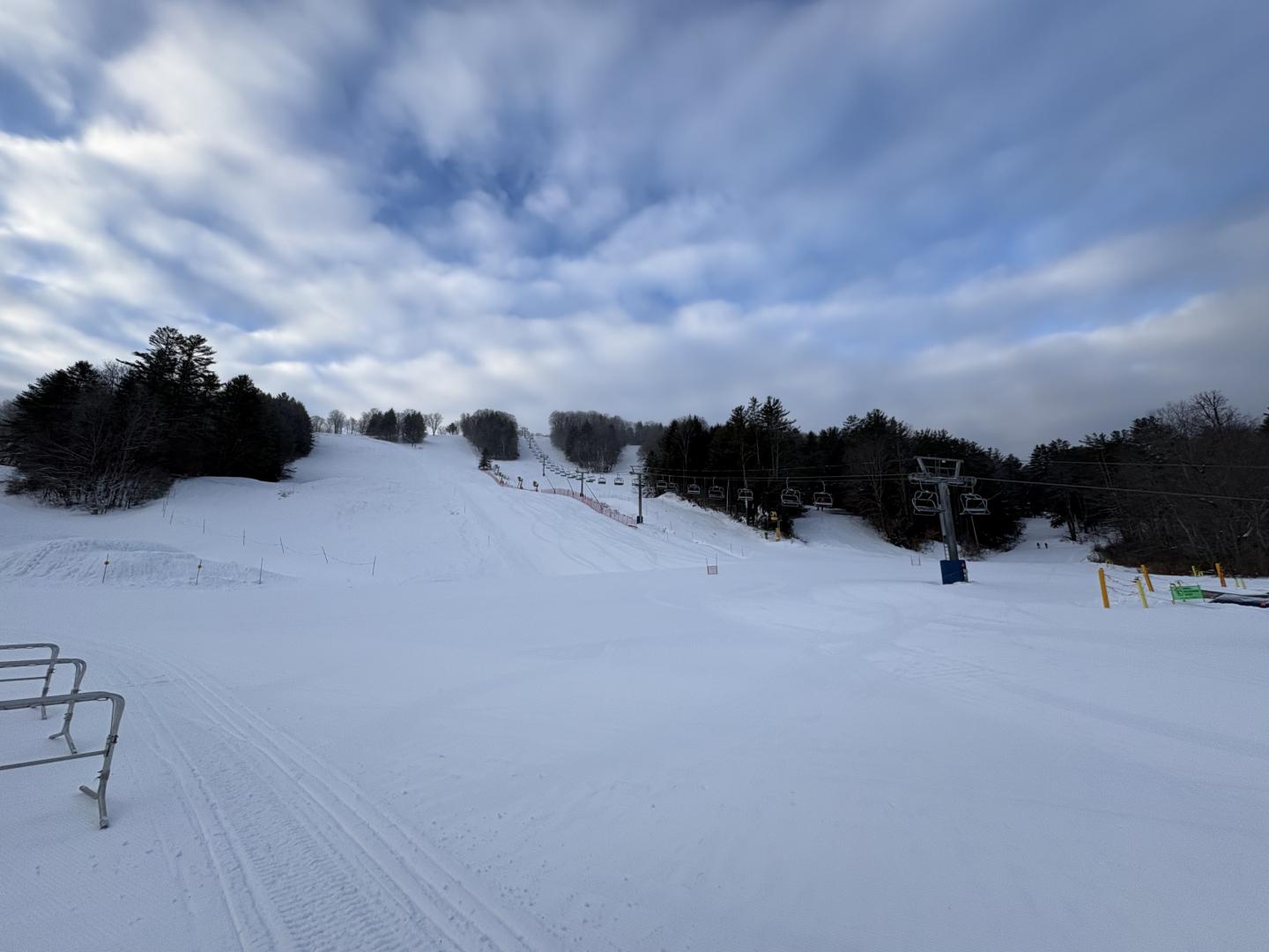 Snow-covered ski slope under a cloudy sky with trees lining the sides.