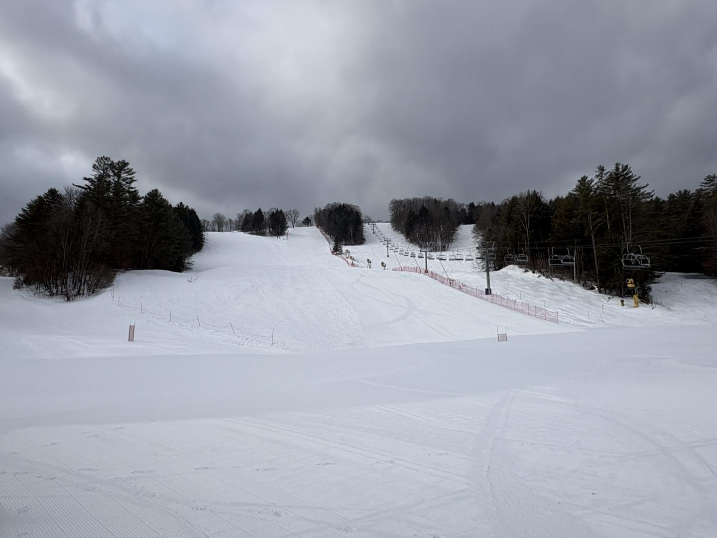 Snow-covered ski slope with cloudy sky and surrounding trees.