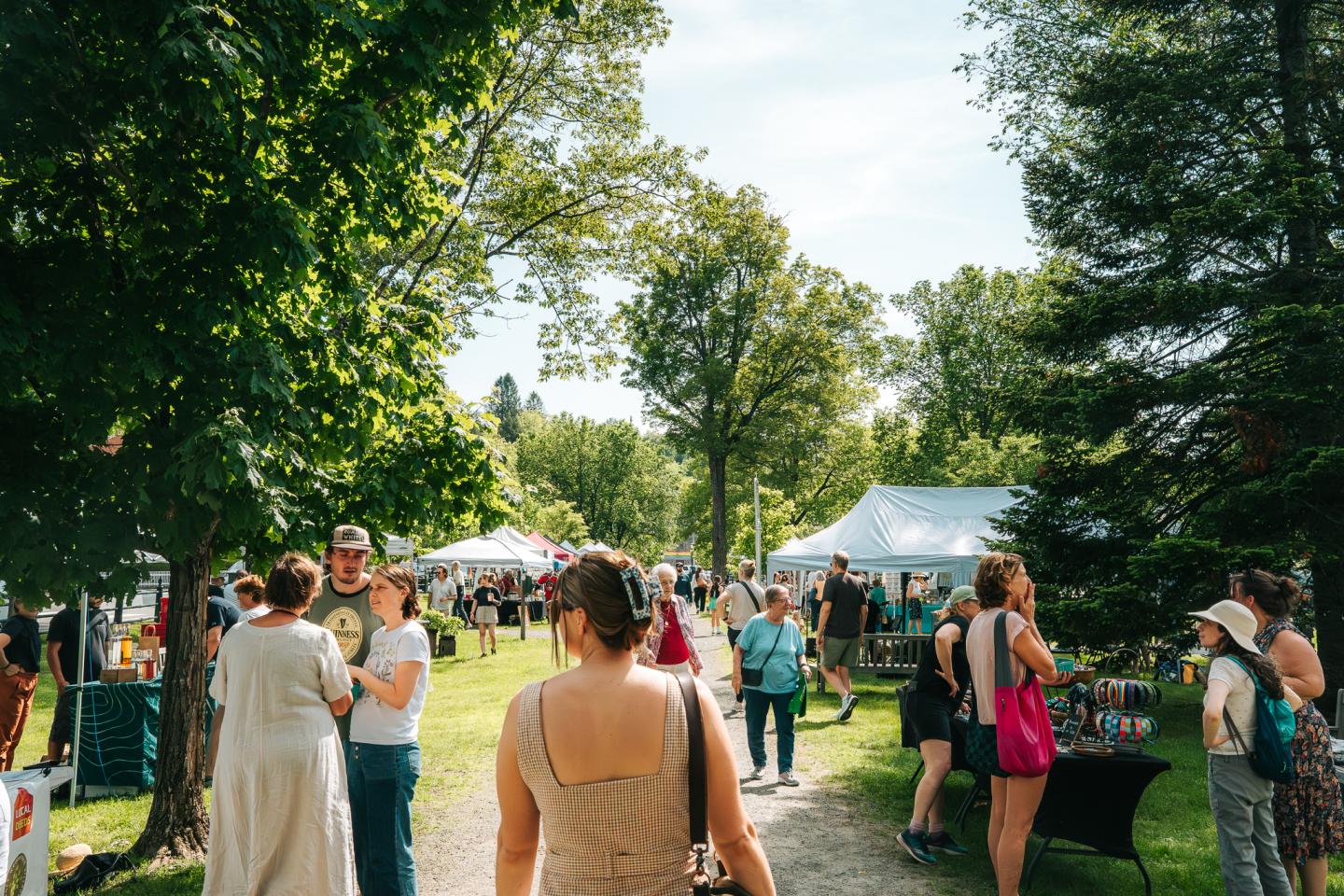 Outdoor market with people walking among trees and white tents on a sunny day.