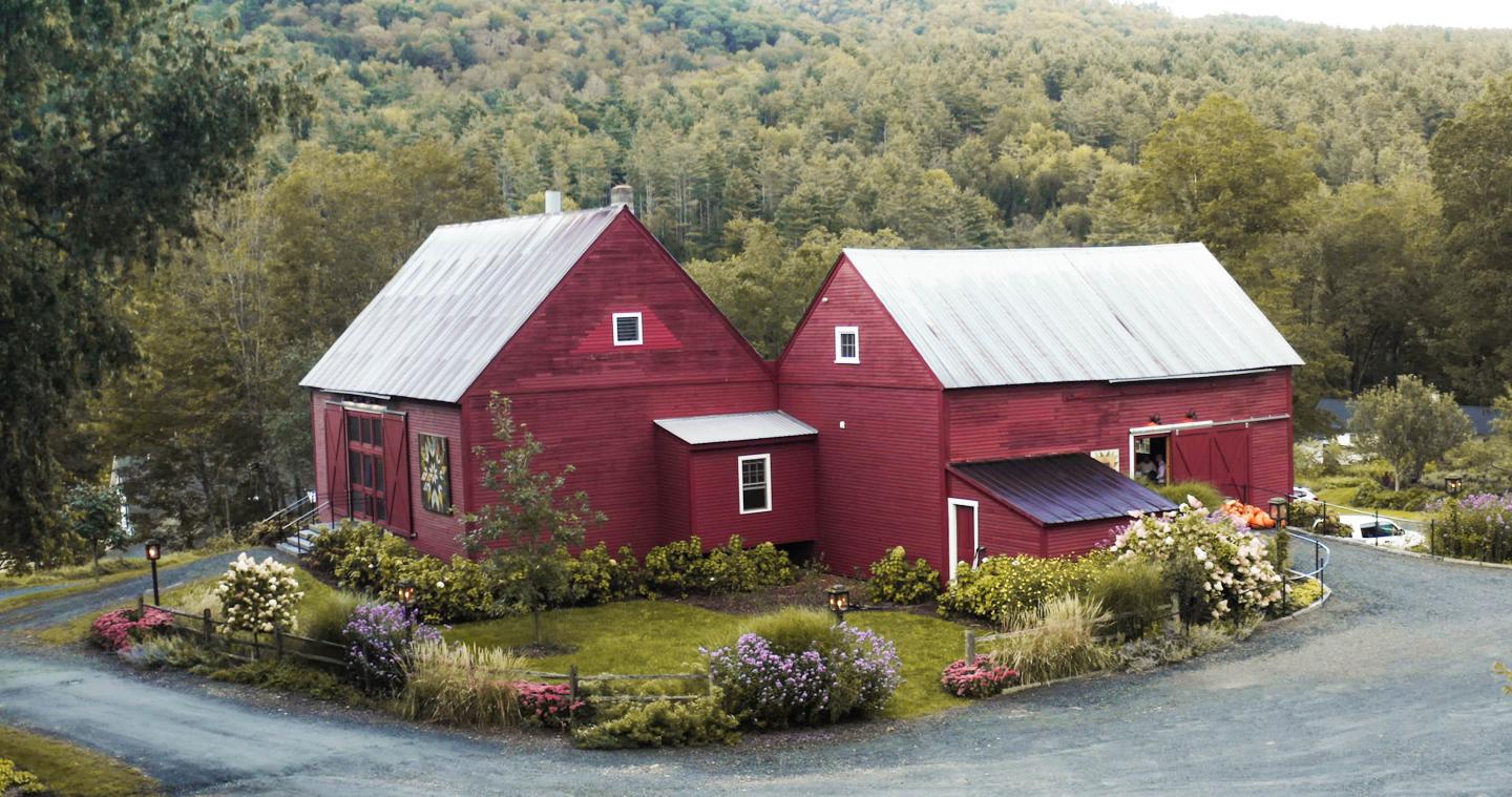 Red barn house with garden, surrounded by trees and gravel path.